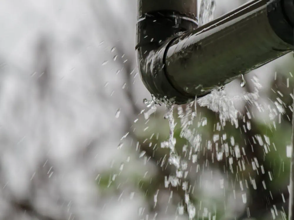 A close-up shot of a leaky metal pipe, possibly a gutter downspout, with water spraying and dripping from a joint or crack. The background is a blurry, light gray.