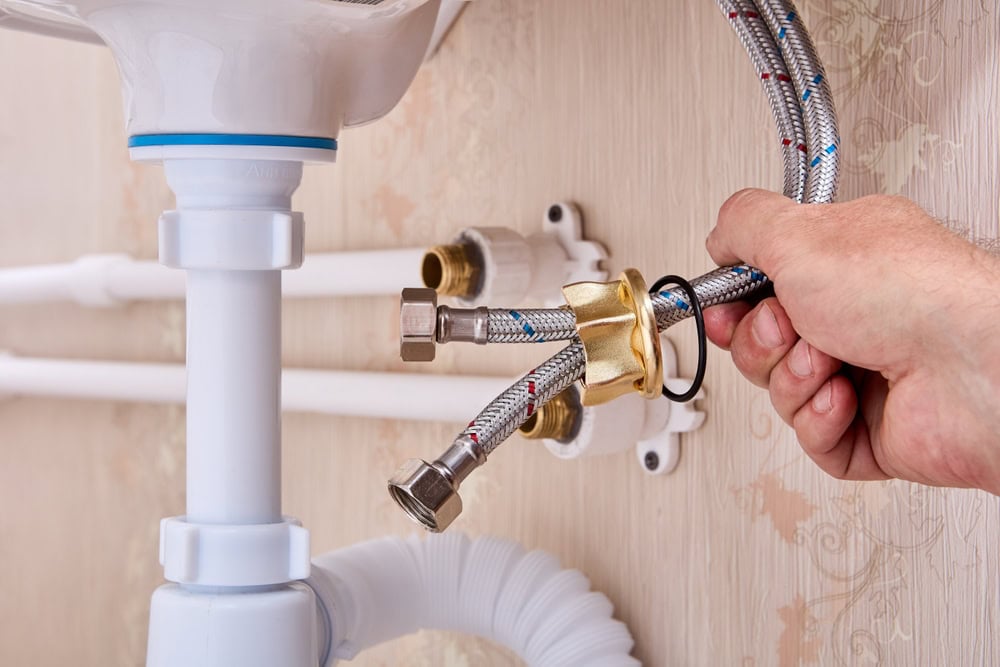 A close-up shot of a person's hand holding and installing flexible braided metal hoses for a sink. The hoses are being connected to brass fittings on the wall, which are attached to white plastic pipes. The wall is a textured beige color.
