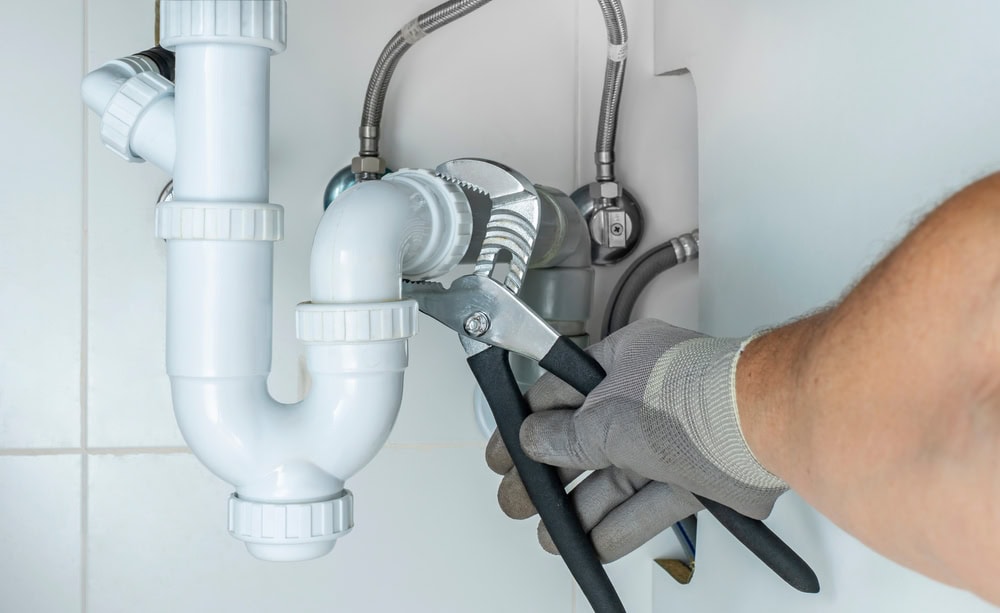 A close-up shot of a person's hand wearing a gray glove, using a pair of large adjustable pliers to tighten a white plastic P-trap pipe under a sink. Braided metal supply lines are visible in the background.