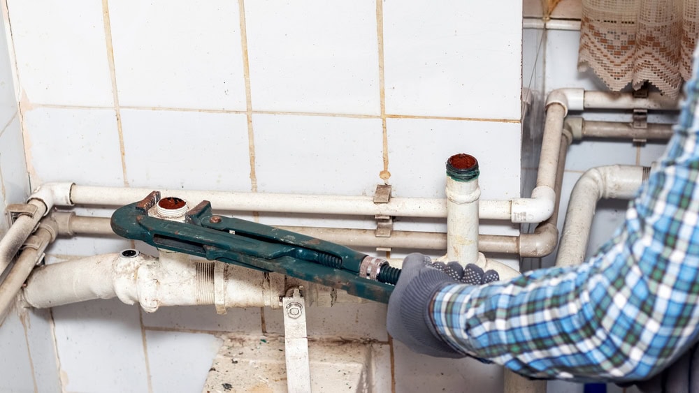 A close-up shot of a person's hands wearing a gray work glove, using a large, dark green pipe wrench to work on white plastic pipes. The pipes are part of a complex plumbing system against a white tiled wall.