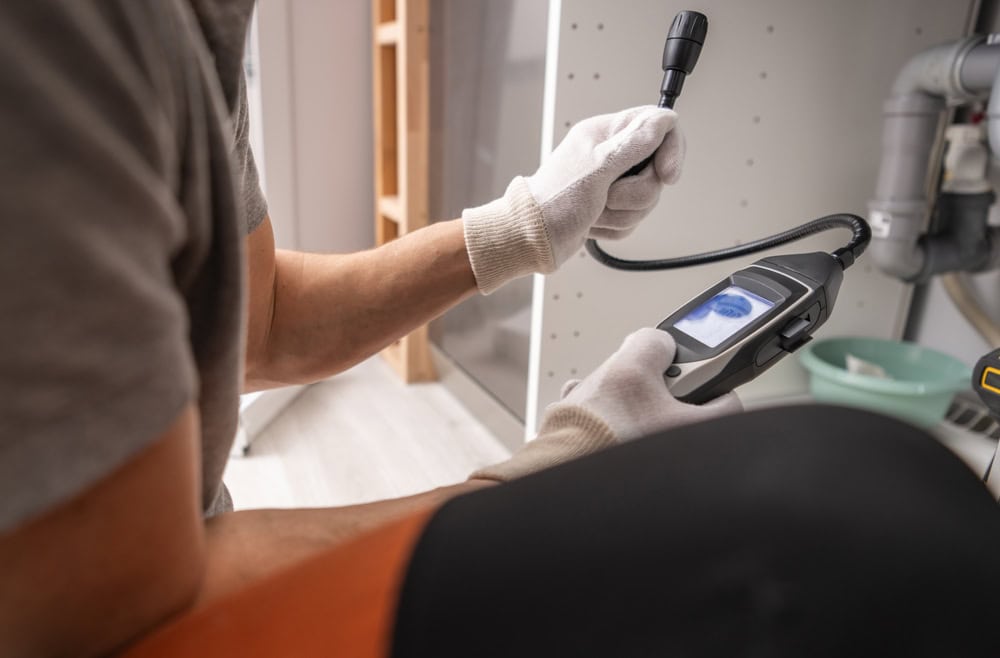 Person wearing gloves uses an inspection camera to examine pipes under a sink. The camera's screen displays a close-up view of the interior. Nearby, plumbing tools and fixtures are visible.