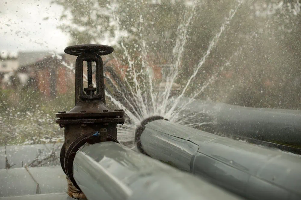 A large water pipe bursts, spraying a powerful jet of water from a broken section near a valve.