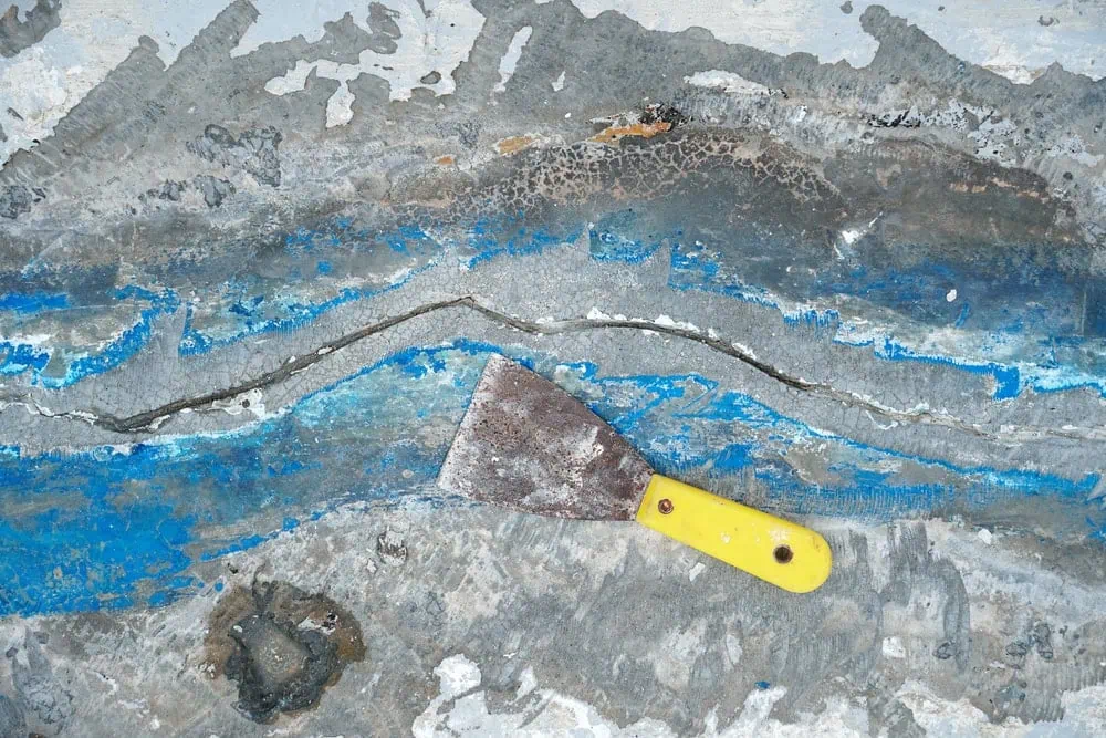 A close-up shot of a cracked concrete surface with a metal scraper tool resting on top of the crack. The surface has patches of blue and gray paint, and the crack is a dark, winding line across the center.