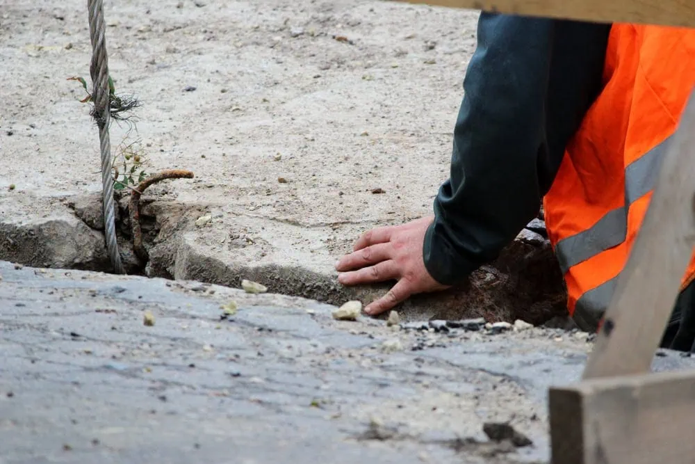 A close-up, low-angle shot of a person's hand touching a jagged crack in a concrete or asphalt surface. The person is wearing a bright orange safety vest. A thick metal cable is visible rising from the crack on the left.