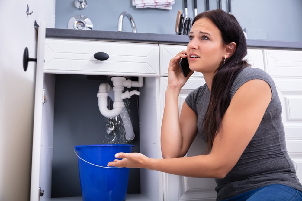 A worried person on the phone holds a blue bucket under leaking pipes beneath a sink in a kitchen, attempting to catch the water.