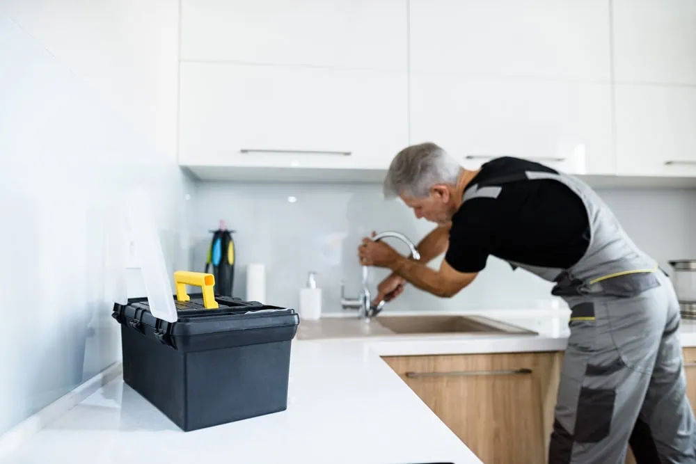 A man with gray hair, wearing overalls, is fixing a kitchen sink faucet. A black toolbox with a yellow handle is on the white countertop. The kitchen has white cabinets and a light wooden finish.
