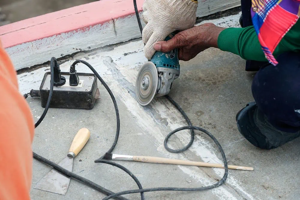 A close-up shot of a person using a small angle grinder with a circular cutting wheel to cut a line into a concrete surface. The person is wearing gloves and is crouched down. An electrical power strip and other tools are visible nearby on the ground.