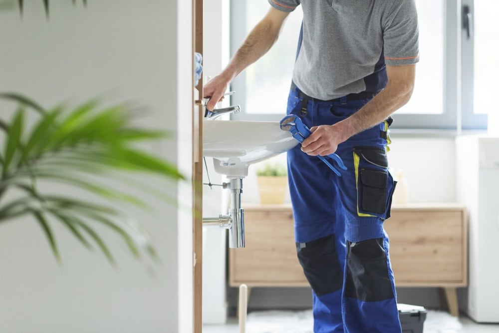 A plumber in blue overalls uses a wrench to fix a white sink in a modern bathroom. There's a wooden cabinet and potted plant in the background, with a large window letting in natural light.