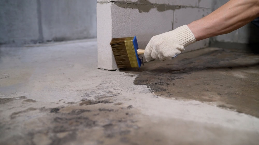 A person's gloved hand holds a large, wide brush, applying a liquid solution to a concrete floor at the base of a concrete block wall. The floor is visibly wet where the solution has been applied.