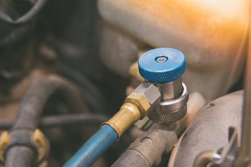 Close-up of a car air conditioning system fitting with a blue valve cap connected to a hose. The background shows parts of the engine bay in soft focus.