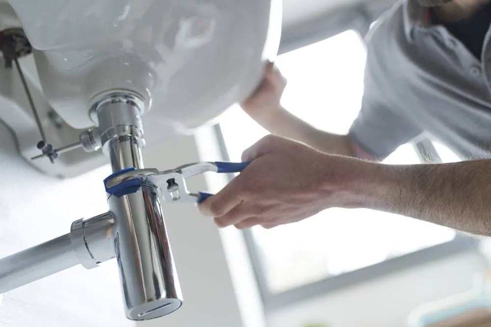 A person uses pliers to tighten a metal pipe under a bathroom sink, performing plumbing maintenance. The scene is well-lit and focused on the hands and plumbing work.