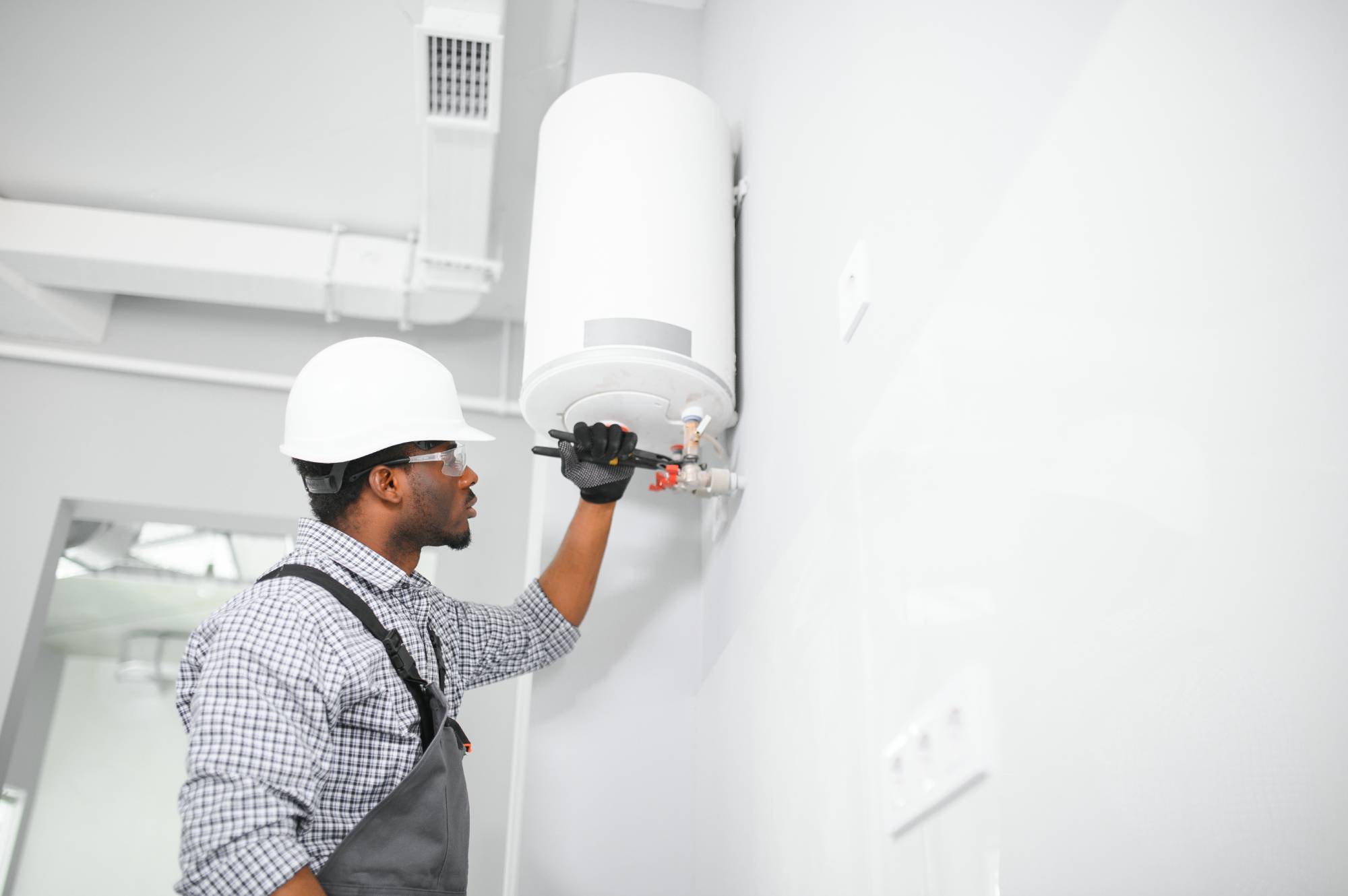 A worker wearing a hard hat, gloves, and overalls uses a screwdriver to repair or install a white water heater mounted on a wall in a bright, modern room.