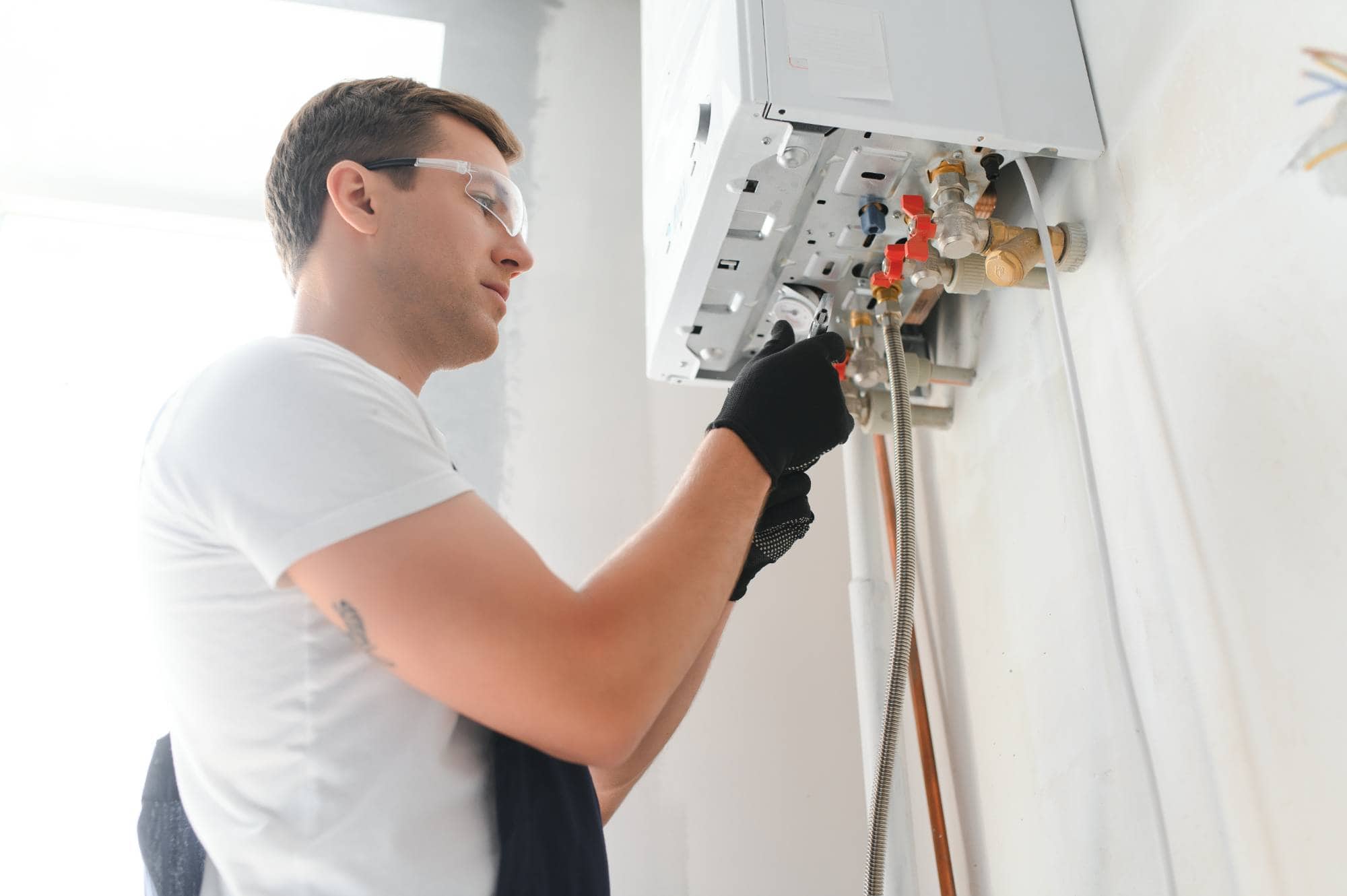 A technician wearing safety glasses and gloves uses tools to repair or install a wall-mounted gas boiler in a bright, indoor setting.