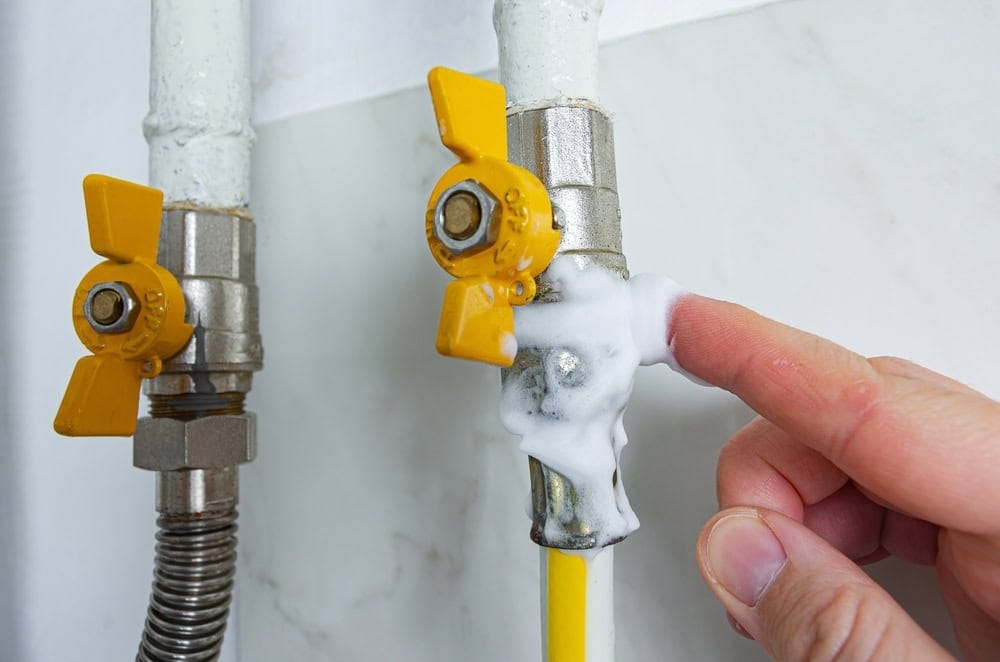 A finger points to a gas valve on a pipe covered in soap bubbles, a test for a leak.