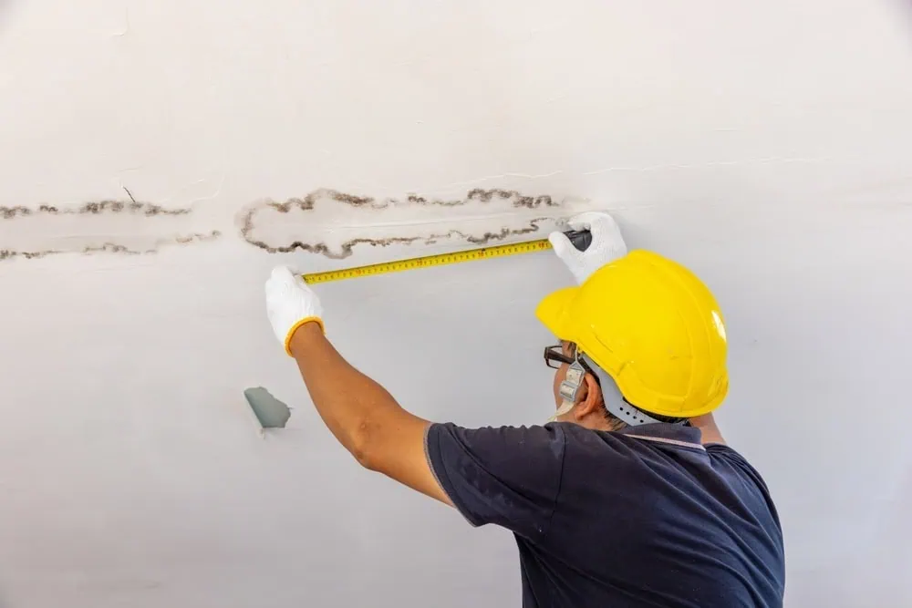 A person in a yellow hard hat and gloves uses a tape measure to assess water damage on a ceiling.