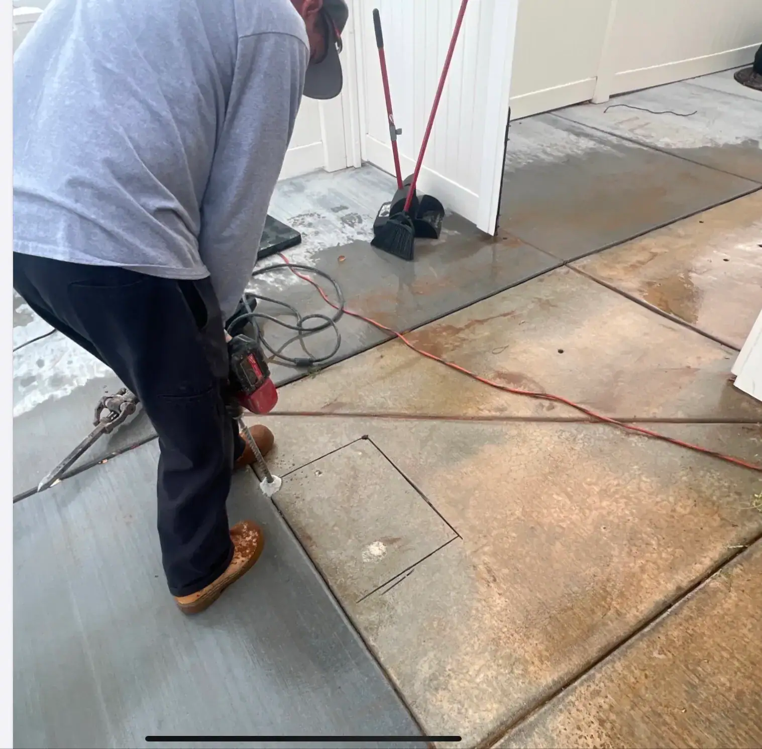 A person wearing a gray long-sleeve shirt, dark pants, and boots uses a power tool to cut a square shape into a concrete surface. Cleaning tools like a broom and dustpan are visible in the background.