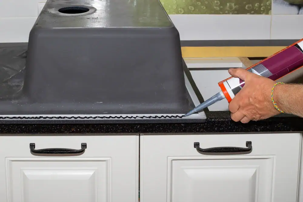 A person applies a bead of sealant with a caulking gun around the edge of an upside-down kitchen sink during installation on a black countertop. White cabinets are visible below the counter.
