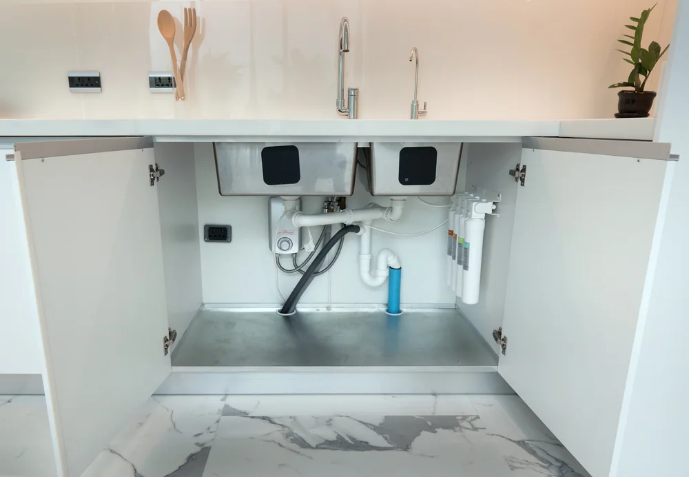 Open kitchen cabinet revealing the plumbing under a sink, including pipes, a water filter system, and a garbage disposal unit. The countertop has minimal items, and the floor is marble-patterned.