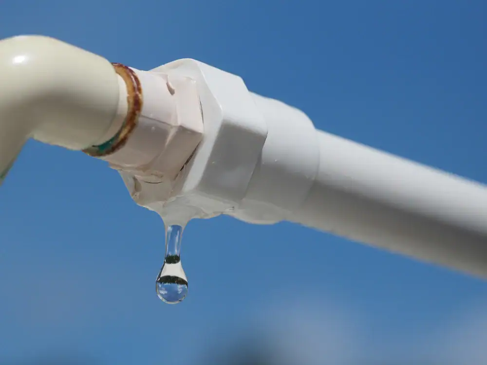 A close-up of a white PVC pipe with a rusted joint, leaking water as a droplet falls against a clear blue sky background.