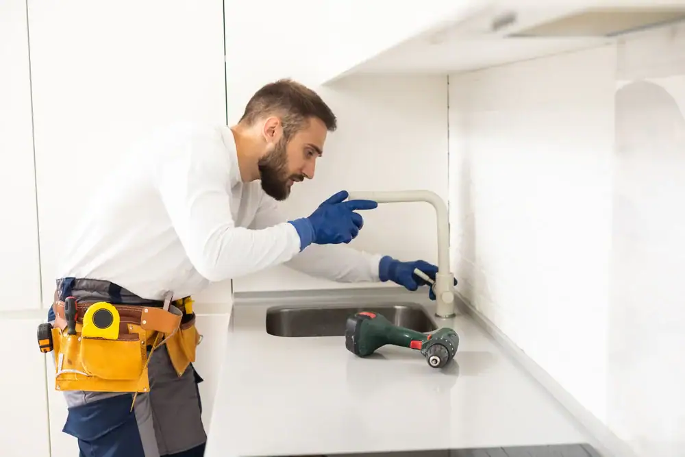 A plumber wearing gloves and a tool belt is installing or repairing a kitchen faucet by the sink, with a cordless drill placed on the countertop.