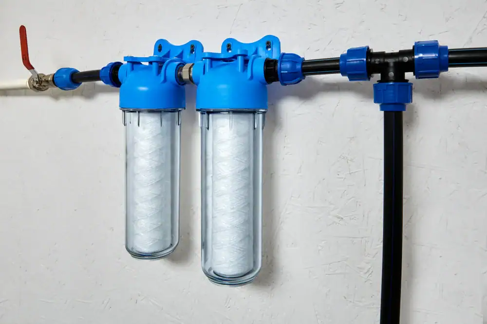 Two blue water filter housings with white cartridges are mounted on a wall, connected to black and white plumbing pipes with various fittings and a red-handled valve.