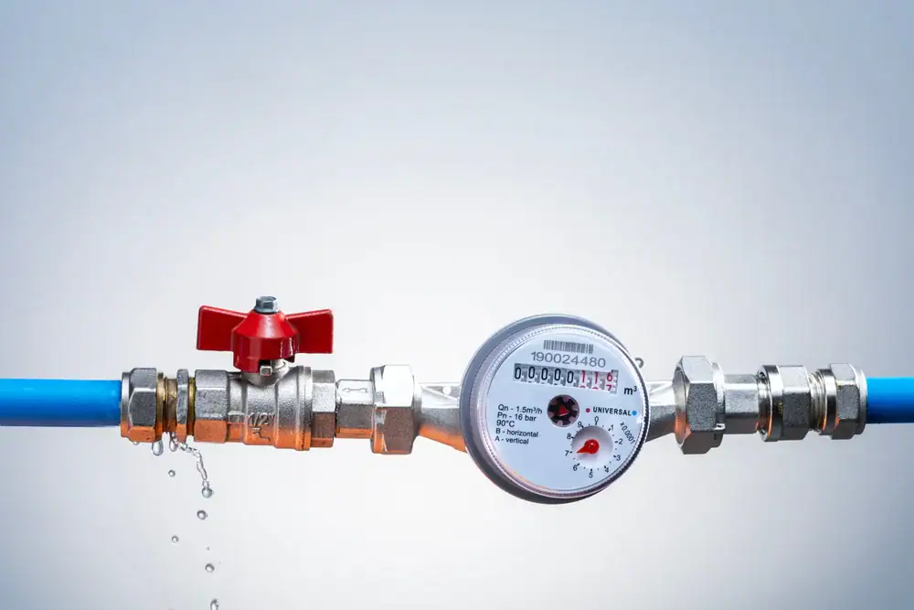 A water meter connected to a blue pipe with a red valve, showing water droplets leaking from a metal joint on the left side against a light gray background.